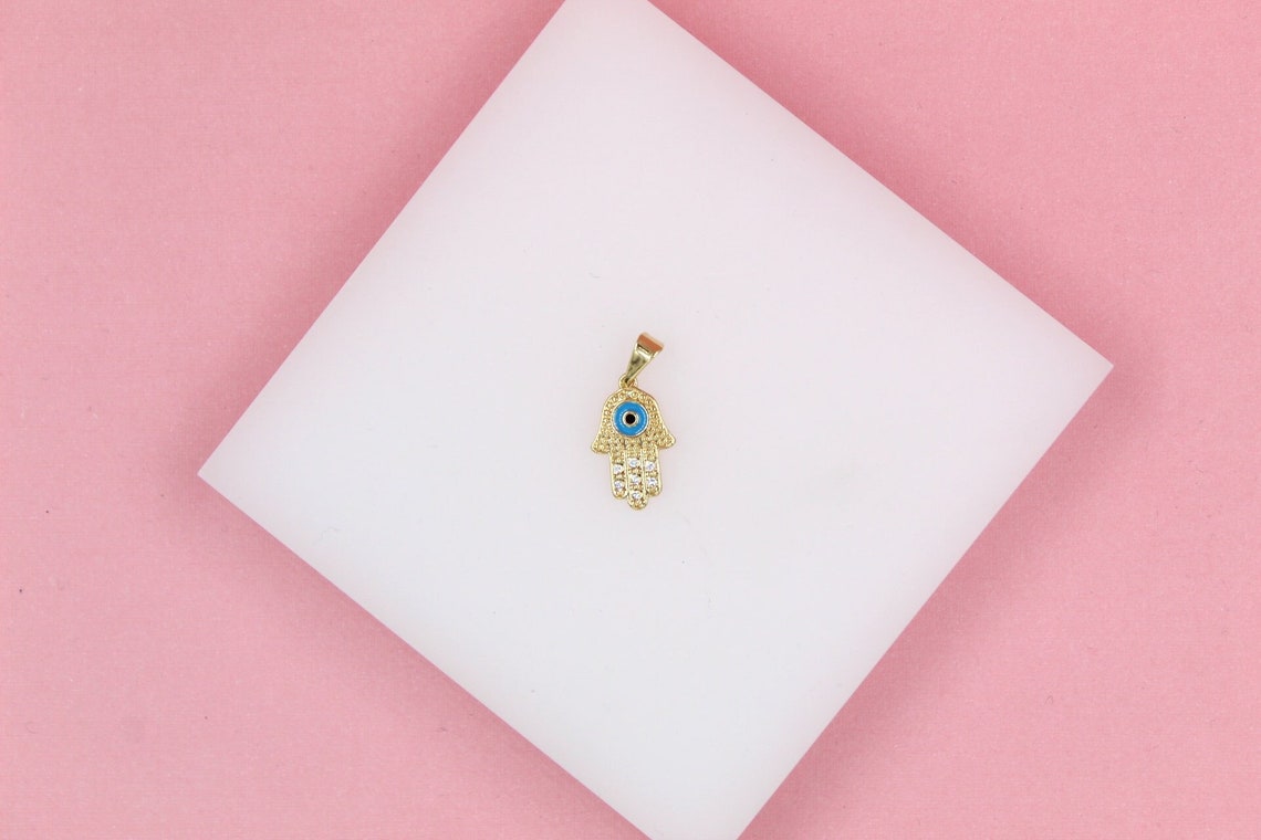 A gold Hamsa hand pendant featuring a blue evil eye and sparkling cubic zirconia stones rests on a white display surface against a pink background.