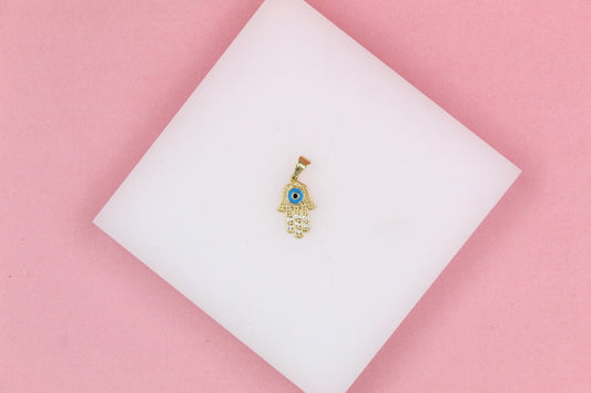 A gold Hamsa hand pendant featuring a blue evil eye and sparkling cubic zirconia stones rests on a white display surface against a pink background.