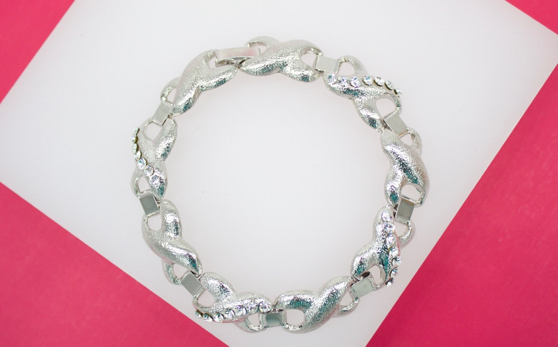A silver link bracelet featuring textured X-shaped charms and sparkling rhinestone accents rests on a white and pink background.