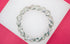 A silver link bracelet featuring textured X-shaped charms and sparkling rhinestone accents rests on a white and pink background.