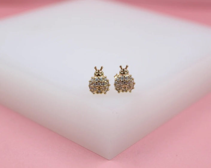 A pair of cute gold ladybug stud earrings encrusted with sparkling cubic zirconia stones rests on a white display surface.