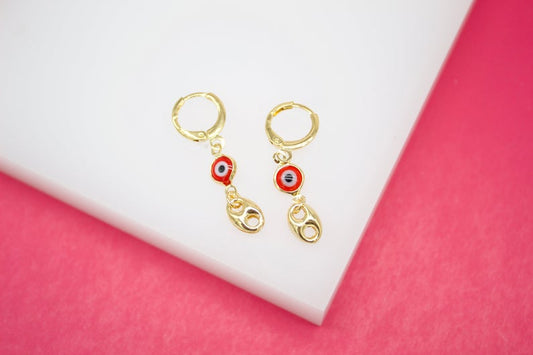A pair of gold huggie hoop earrings features hanging red evil eye charms and interlocking link pendants resting on a white surface against a pink background.