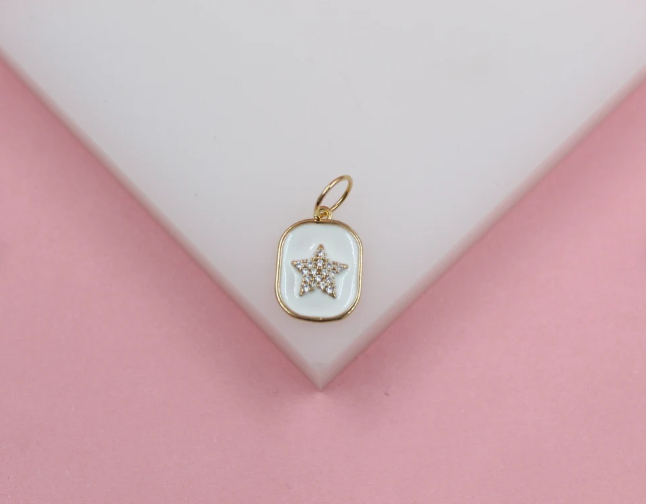 A gold rectangular pendant featuring a white enamel background and a sparkling rhinestone star rests on a white display against a pink backdrop.