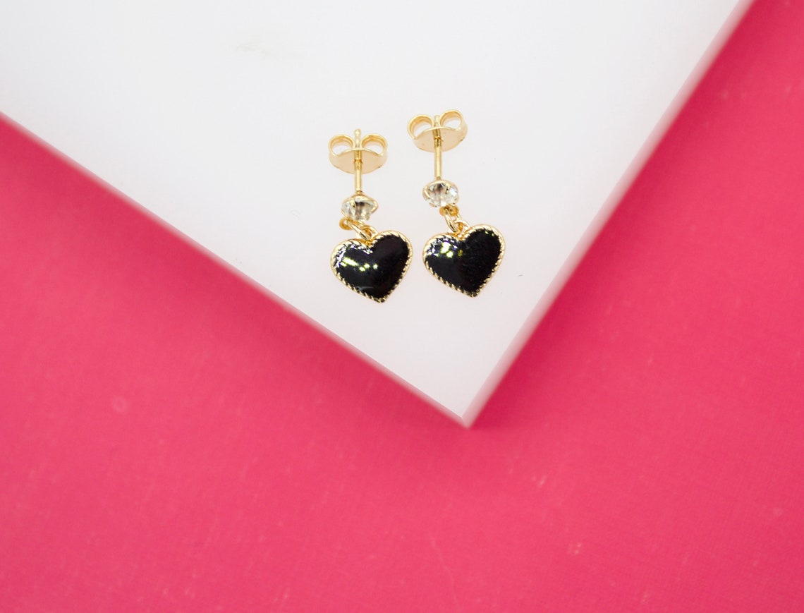 A pair of gold stud earrings featuring dangling black enamel heart charms rests on a white display surface against a vibrant pink background.