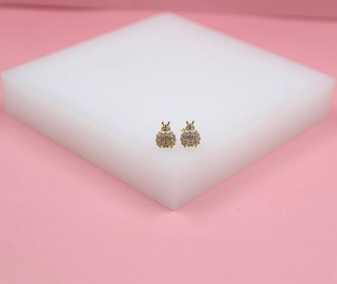 A pair of sparkling ladybug stud earrings rests on a white display block against a soft pink background.
