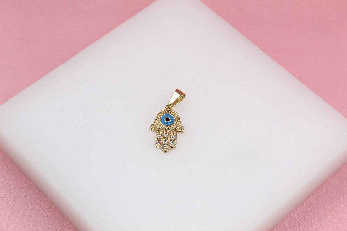 A gold Hamsa hand pendant featuring a blue evil eye and sparkling crystal accents rests on a white display surface against a pink background.