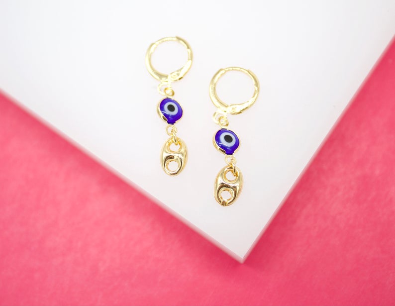 A pair of gold huggie hoop earrings featuring dangling blue evil eye charms and interlocking links rests on a white display against a pink background.