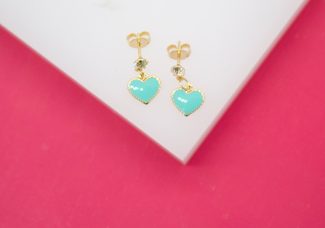 A pair of gold heart-shaped stud earrings featuring vibrant turquoise enamel rests on a white display surface against a vibrant pink background.