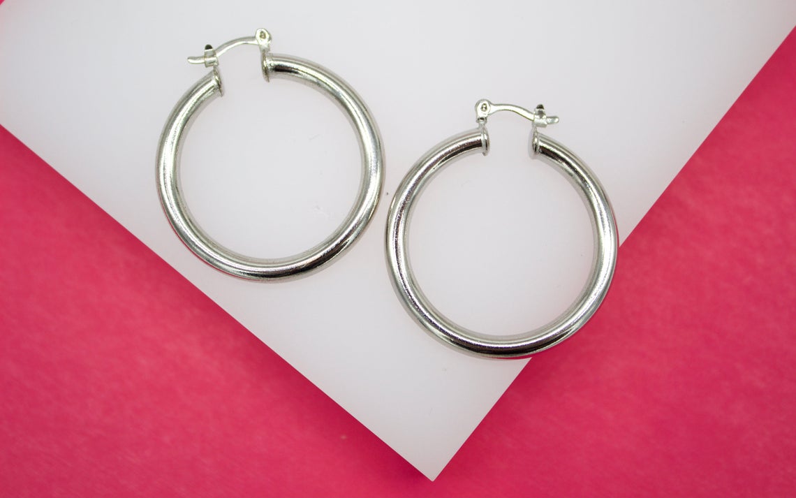 A pair of polished silver hoop earrings rests on a white display surface against a vibrant pink background.