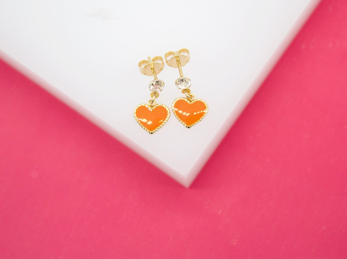 A pair of gold earrings featuring sparkling crystal studs and bright orange enamel heart charms rests on a white display against a vibrant pink background.