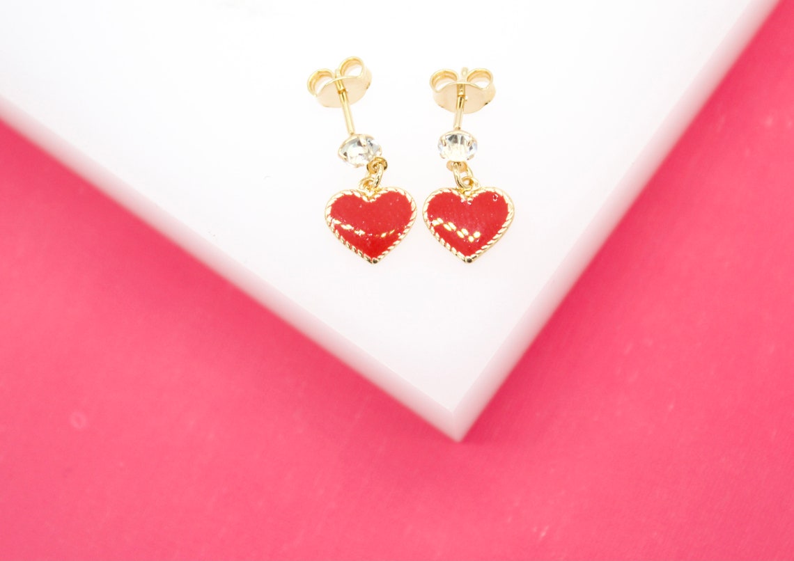 A pair of gold heart-shaped earrings featuring red enamel and small crystals rests on a white display surface against a vibrant pink background.