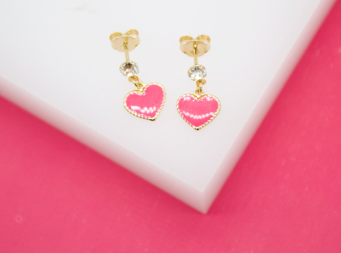 A pair of gold earrings featuring glittering crystal studs and dangling hot pink heart charms resting on a white display against a vibrant pink background.
