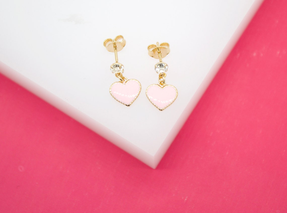A pair of gold dangle earrings featuring sparkling cubic zirconia crystals and charming pink enamel hearts rest on a white surface against a bright pink background.