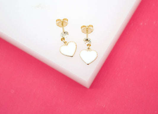A pair of delicate gold heart-shaped stud earrings featuring white enamel and sparkling crystal accents rest on a white display block against a vibrant pink background.