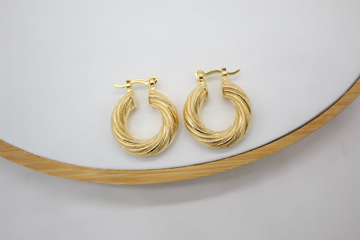 A pair of textured gold hoop earrings rests on a white display surface with a wooden trim.