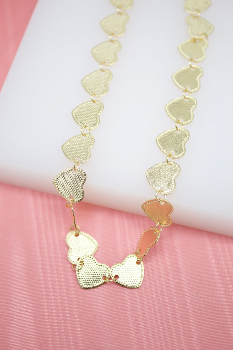 A gold heart-link necklace rests gracefully on a white display against a pink background.