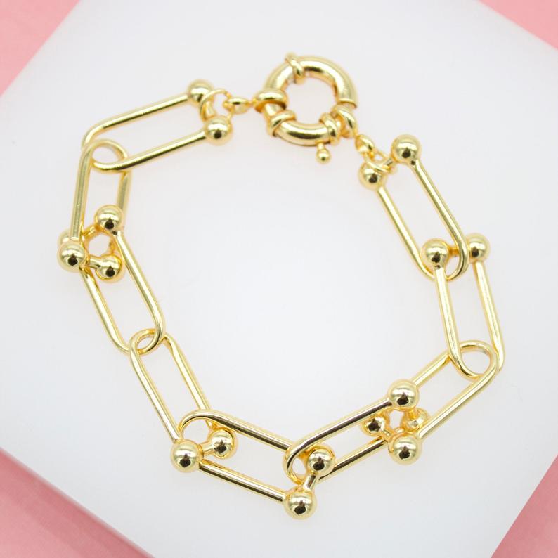 A gold paperclip chain bracelet featuring alternating bead details rests on a white display surface.