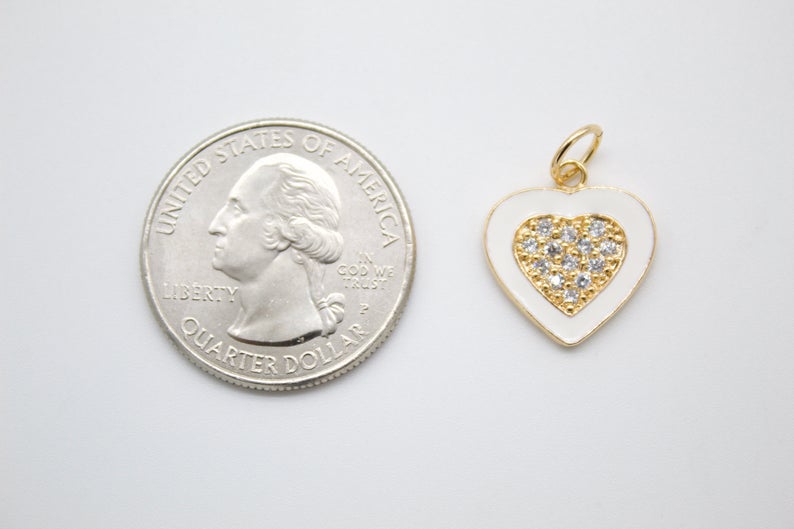 A gold-plated white enamel heart charm featuring a paved cubic zirconia center rests next to a US quarter coin for scale.