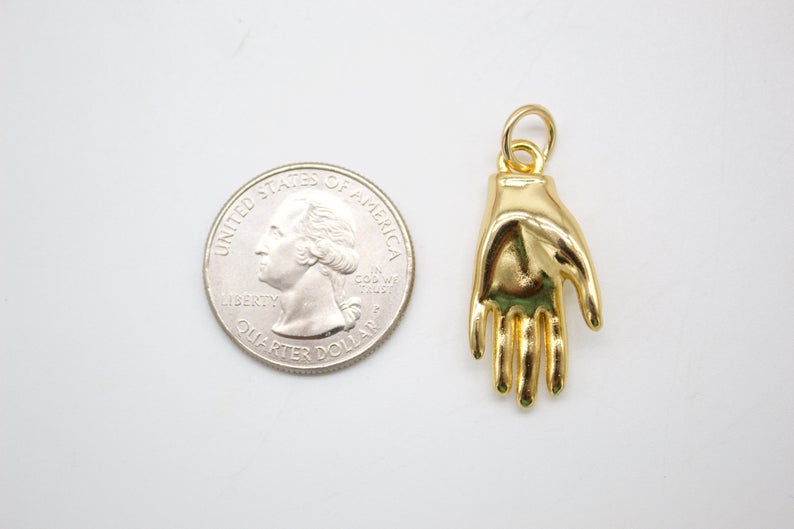 A shiny gold hand charm rests next to a US quarter coin for size comparison on a white surface.