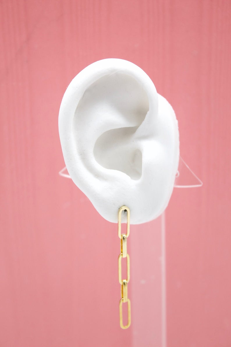 A white mannequin ear displays a dangling gold paperclip chain earring against a pink background.