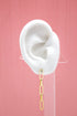 A white mannequin ear displays a dangling gold paperclip chain earring against a pink background.