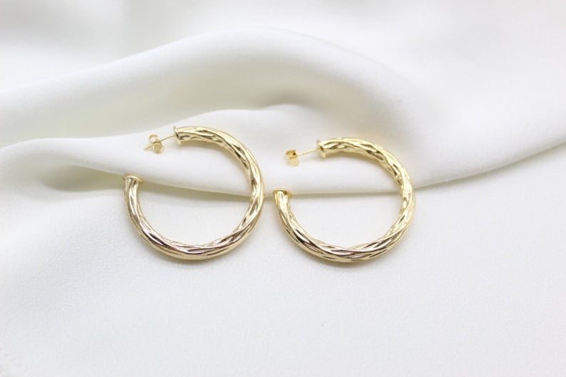 A pair of textured gold hoop earrings rests gracefully on a white silk backdrop.