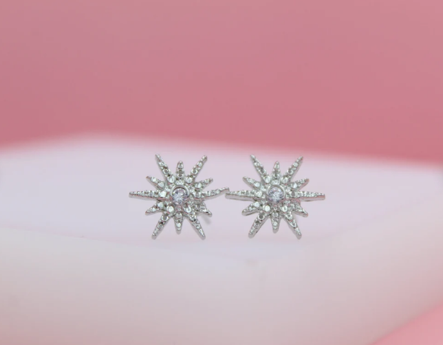 A pair of sparkling starburst stud earrings rests on a soft pink background.