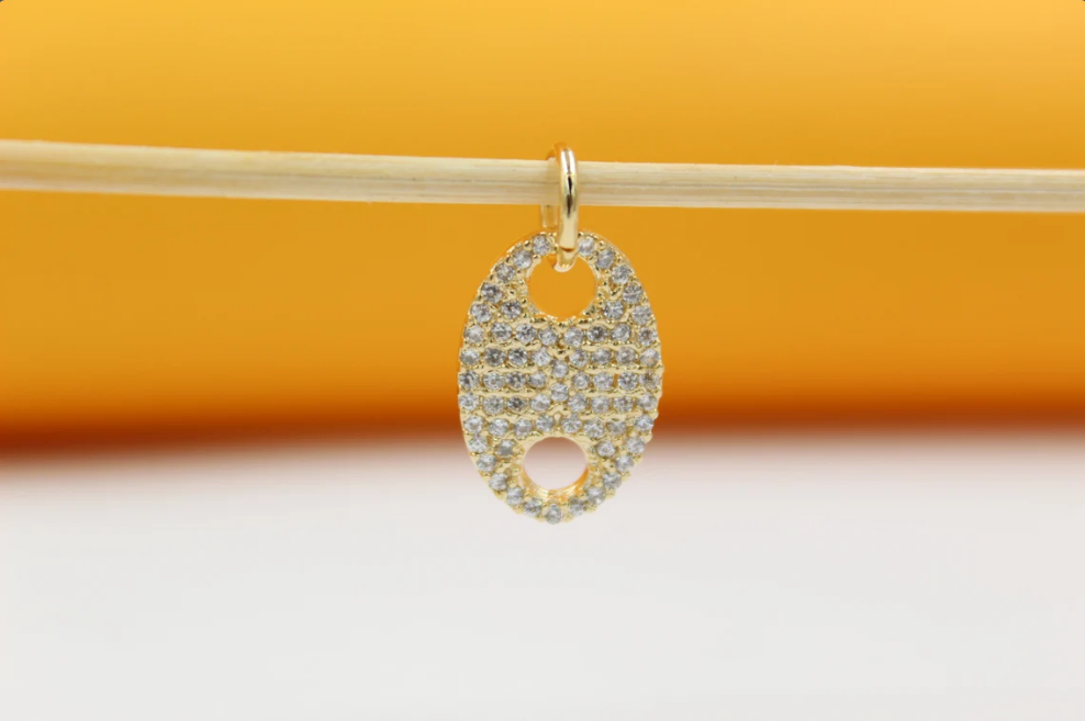 A gold-plated oval pendant encrusted with sparkling cubic zirconia stones hangs gracefully from a thin wooden stick against a warm yellow background.