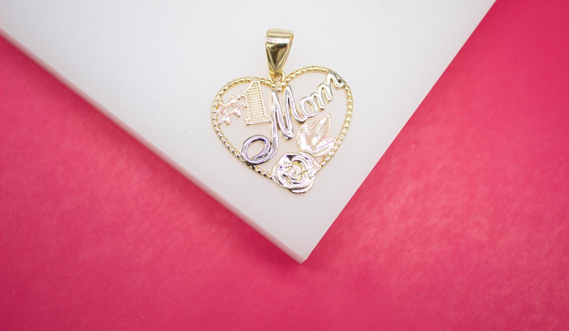 A gold and silver heart-shaped pendant inscribed with #1 Mom rests on a white display against a vibrant pink background.