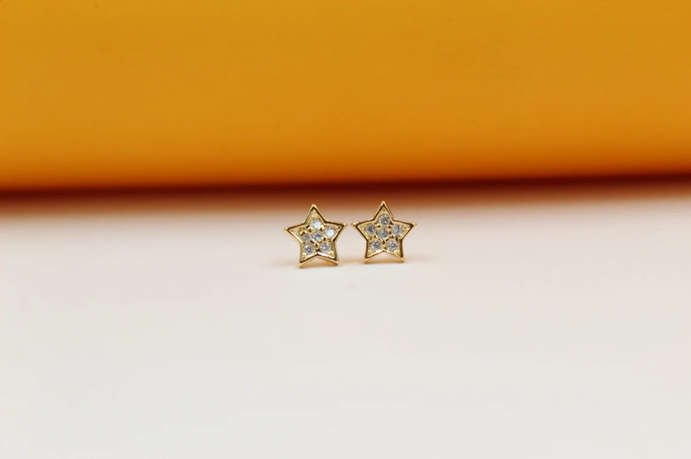 A pair of gold star-shaped stud earrings featuring embedded cubic zirconia stones rests on a neutral surface against an orange backdrop.