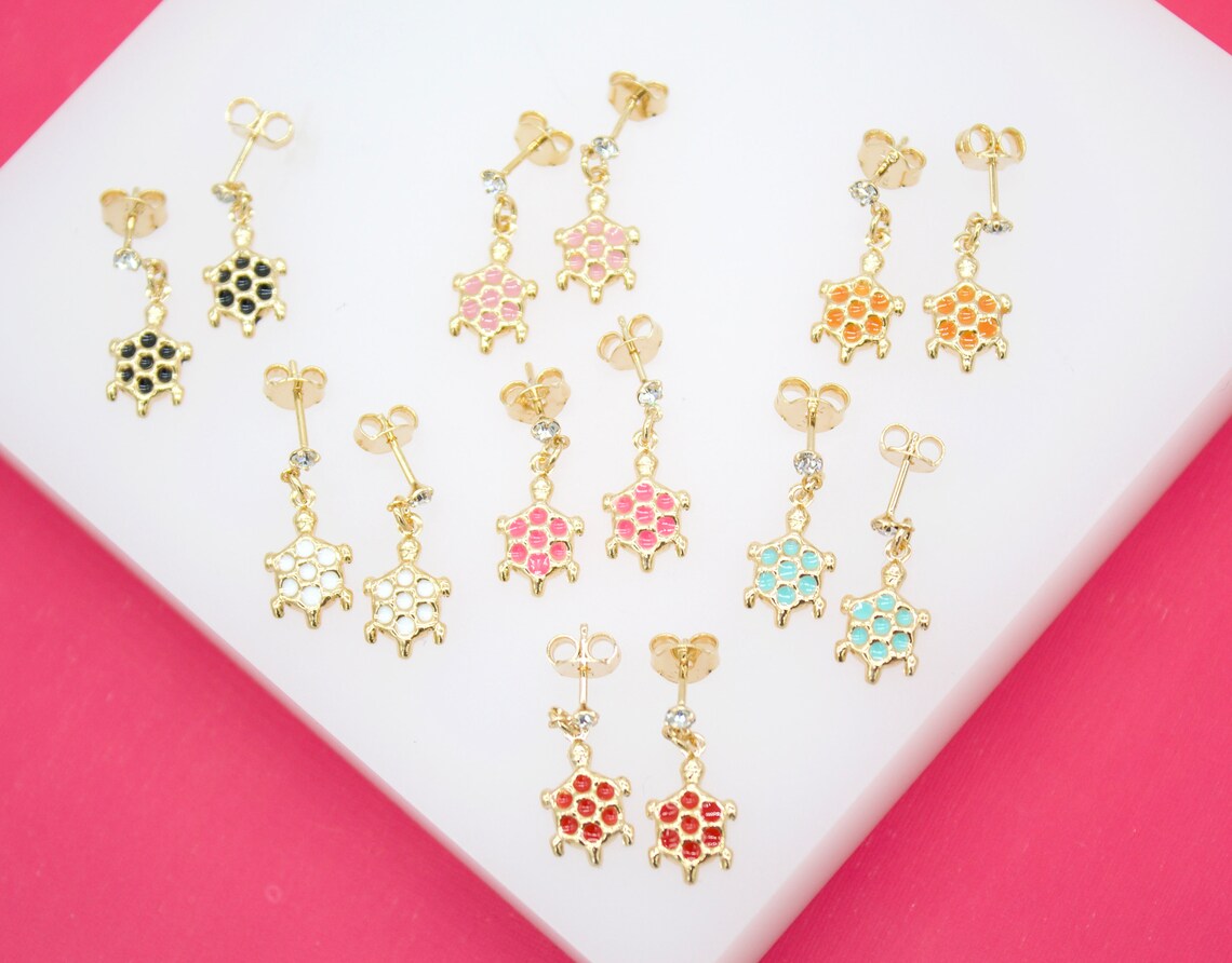 Several pairs of gold turtle earrings with colorful enamel shells are arranged neatly on a white display surface against a vibrant pink background.