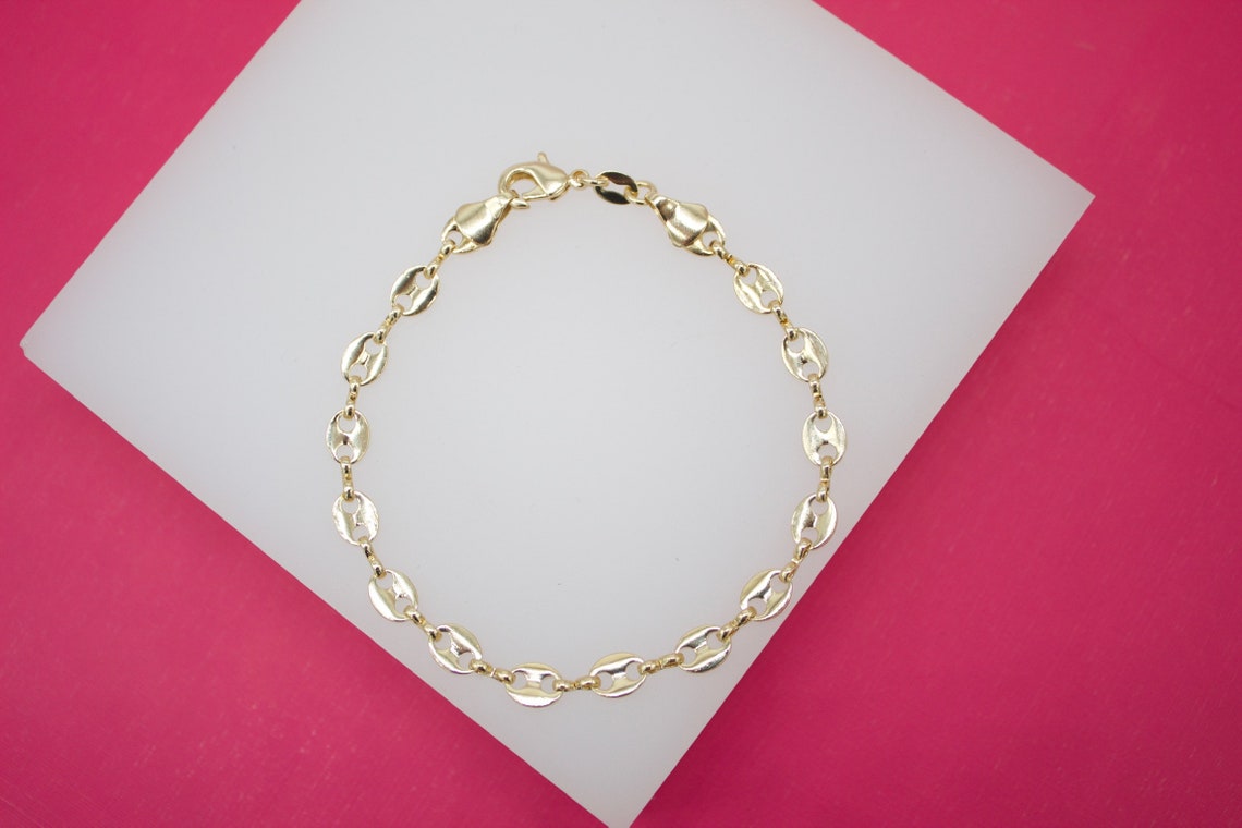 A delicate gold coffee bean chain bracelet rests on a white display against a vibrant pink background.