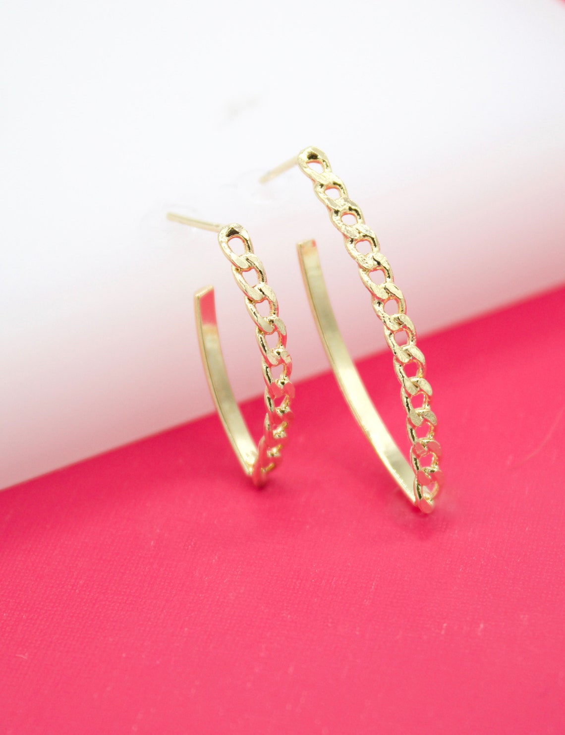 A pair of gold chain-link hoop earrings rests against a white and pink background.