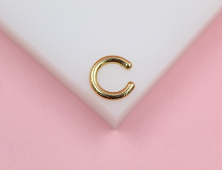A small gold initial C ear cuff rests on a white display against a pink background.