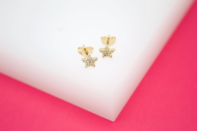 Two gold star-shaped stud earrings sparkle with pave diamonds against a white and pink background.