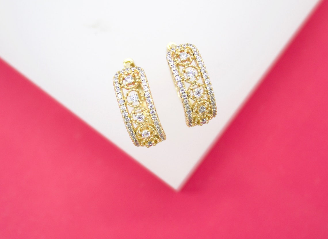A pair of ornate gold hoop earrings featuring intricate filigree work and sparkling cubic zirconia stones rests on a white display against a vibrant pink background.