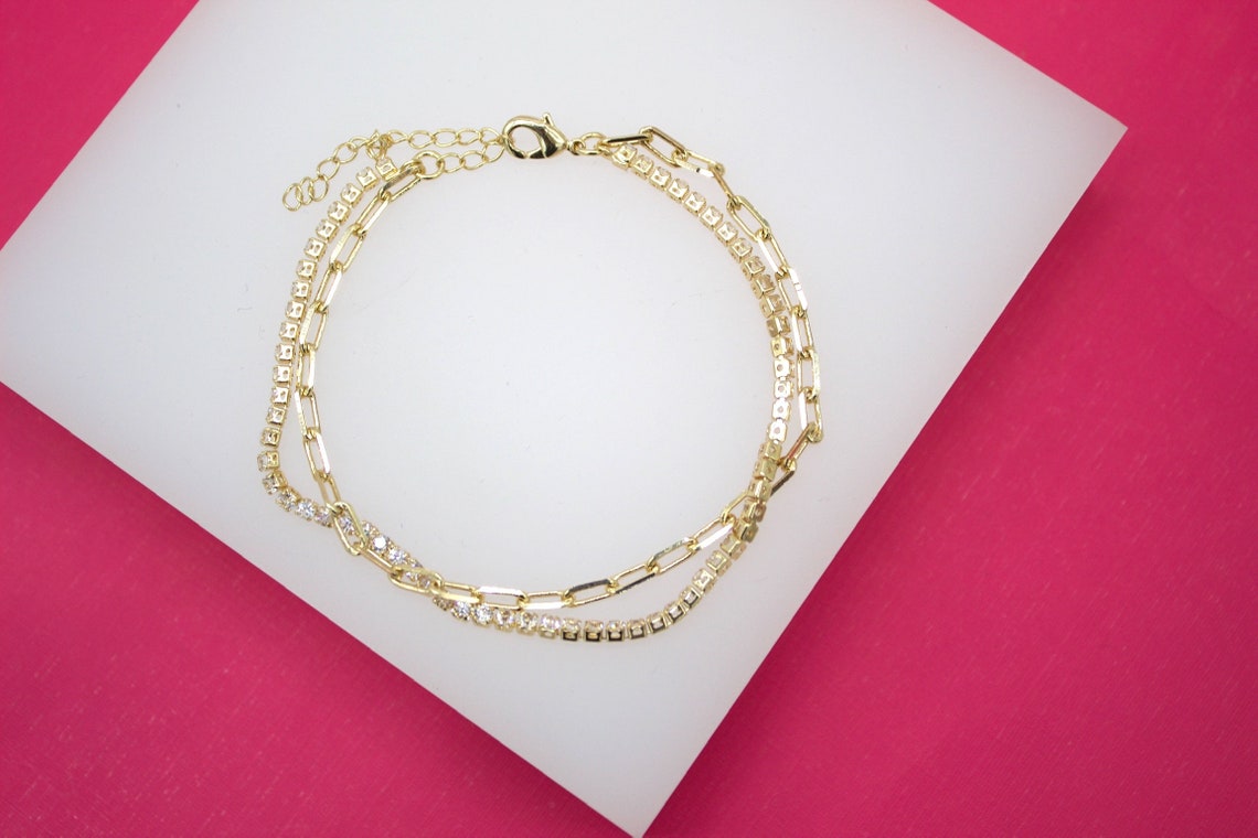 A double-strand gold bracelet featuring a delicate link chain and a sparkling rhinestone tennis chain rests on a white display against a vibrant pink background.