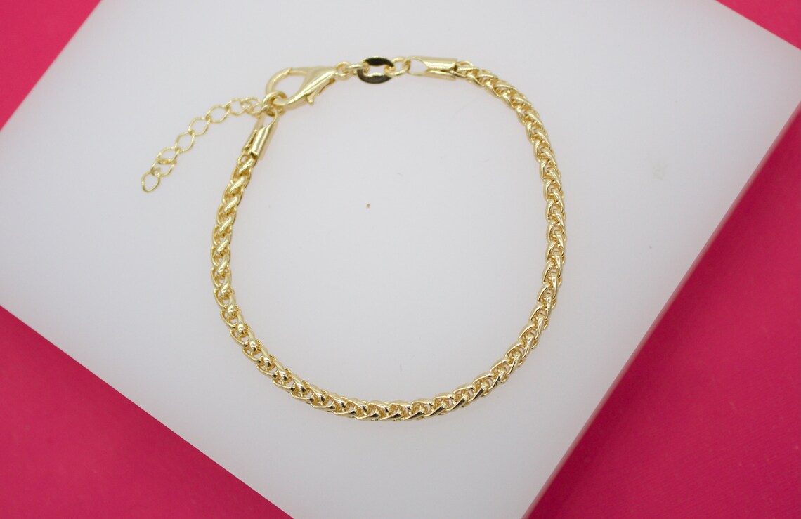 A delicate gold wheat chain bracelet rests on a white display surface against a vibrant pink background.