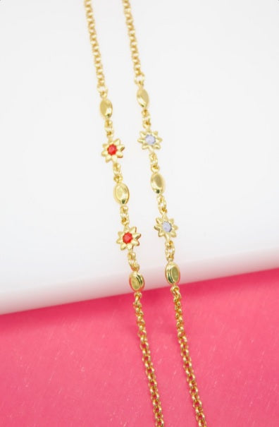 A delicate gold chain necklace features alternating red and white crystal flower stations against a bright pink and white background.