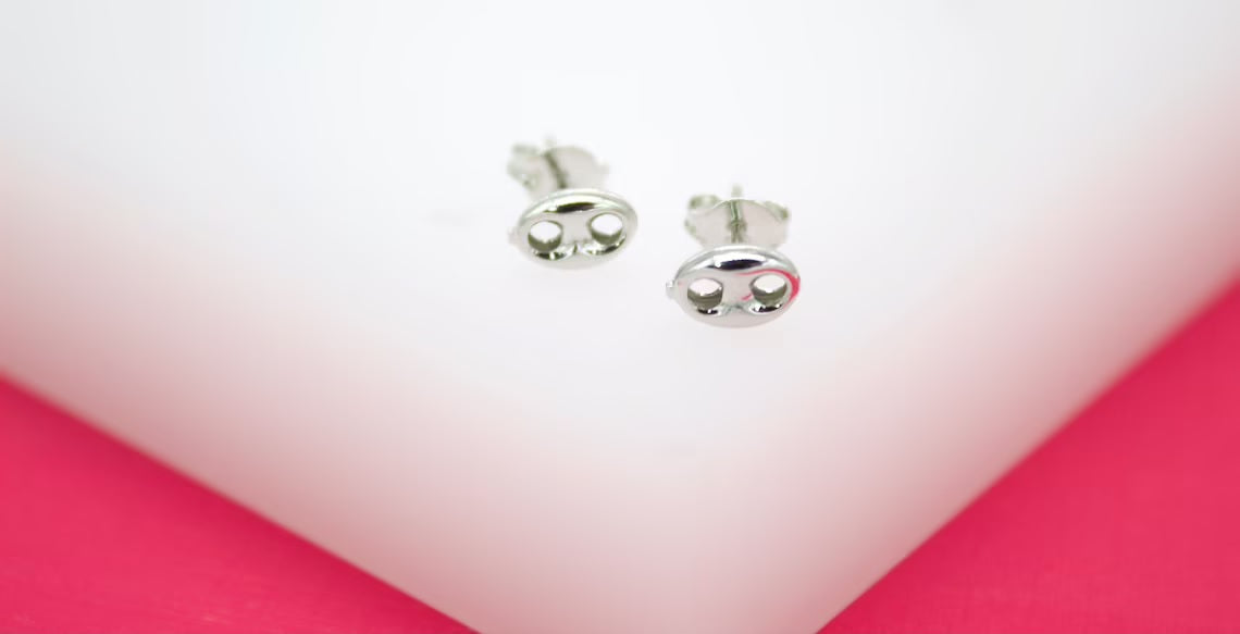 Two silver coffee bean stud earrings rest on a clean white surface against a bright pink background.