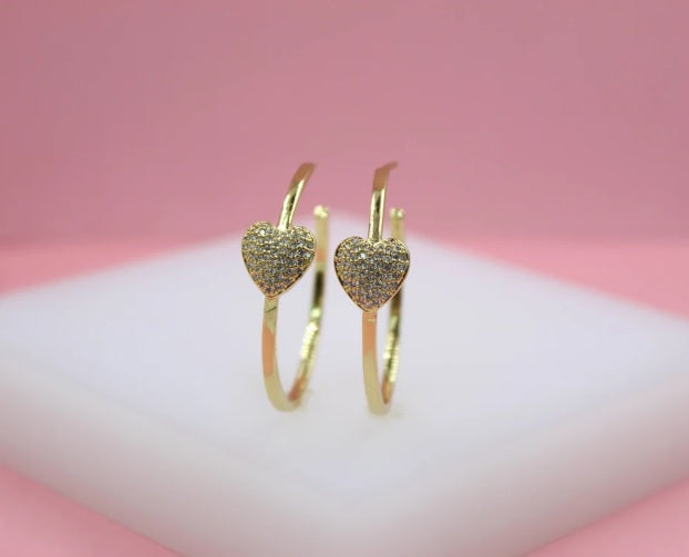A pair of gold hoop earrings featuring sparkling pavé diamond hearts displayed upright on a white platform.