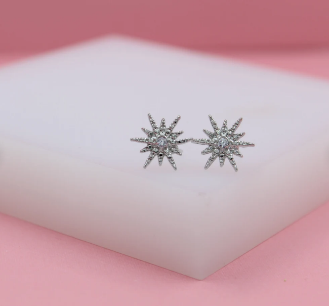 Two sparkly starburst silver stud earrings rest on a white display block against a soft pink backdrop.
