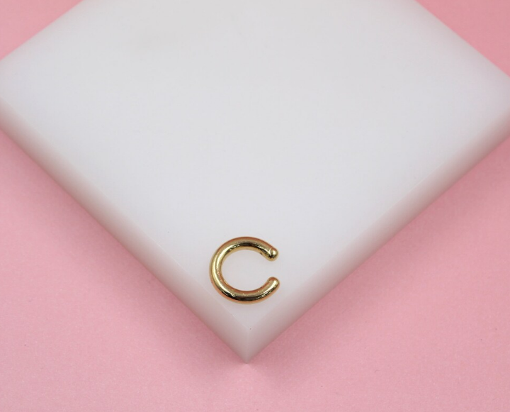 A shiny gold initial C ear cuff rests on a white display block against a soft pink background.