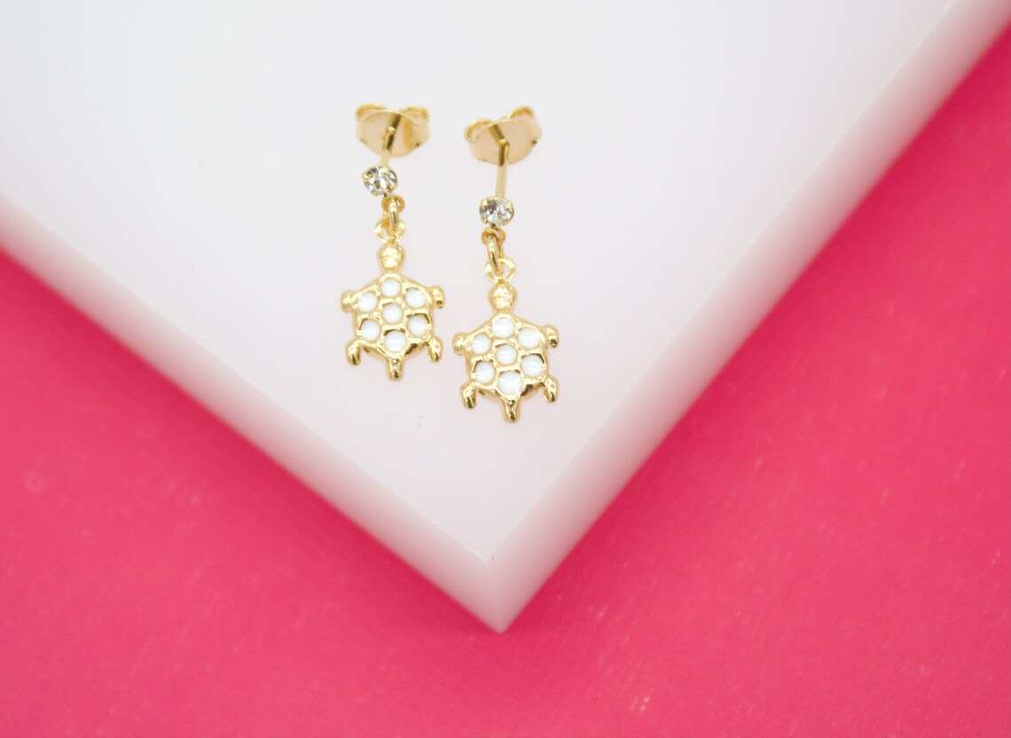 A pair of gold turtle stud earrings featuring white enamel shell details and sparkling cubic zirconia stones rests on a white display against a vibrant pink background.