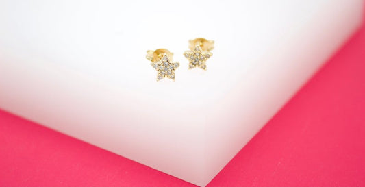 A pair of gold star stud earrings encrusted with tiny diamonds rests on a white and pink surface.