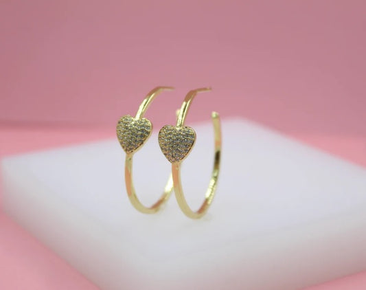 A pair of gold hoop earrings featuring sparkling pavé diamond hearts rests on a white display against a pink backdrop.
