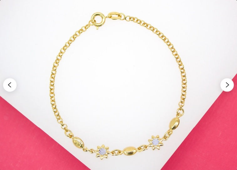 A delicate gold chain bracelet features alternating flower charms with purple gemstones and smooth oval beads, displayed against a pink and white background.