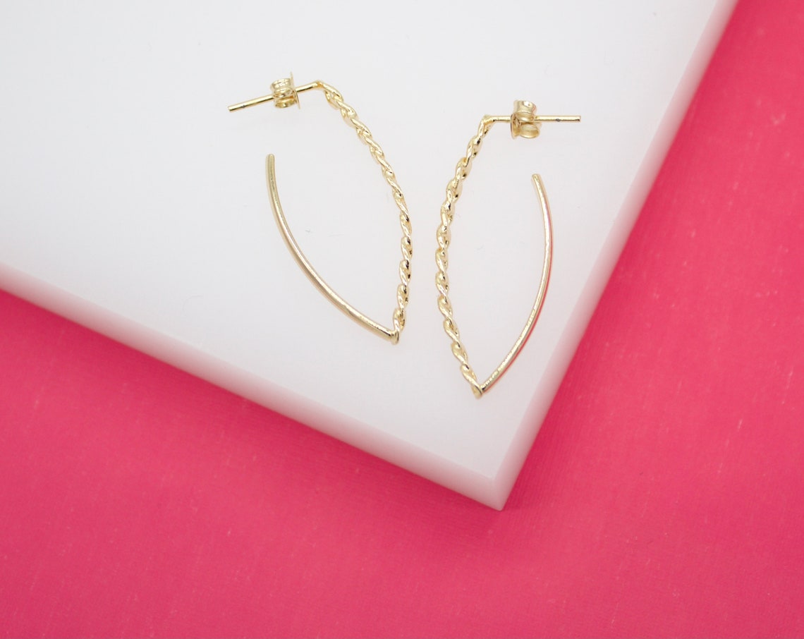 A pair of elegant twisted gold hoop earrings rests on a white block against a vibrant pink background.