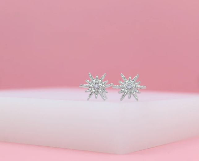 Sparkling silver starburst stud earrings rest on a white display against a soft pink backdrop.