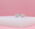 Sparkling silver starburst stud earrings rest on a white display against a soft pink backdrop.